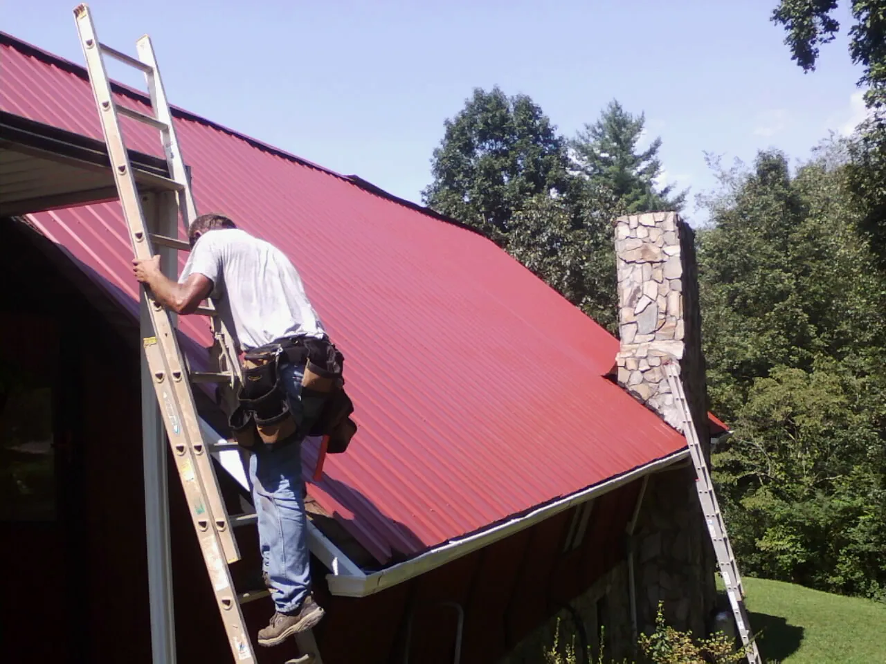 Roofer on a ladder beside a red metal roof and stone chimney — Soffit & Fascia Repair by Roofing Brothers in Moline